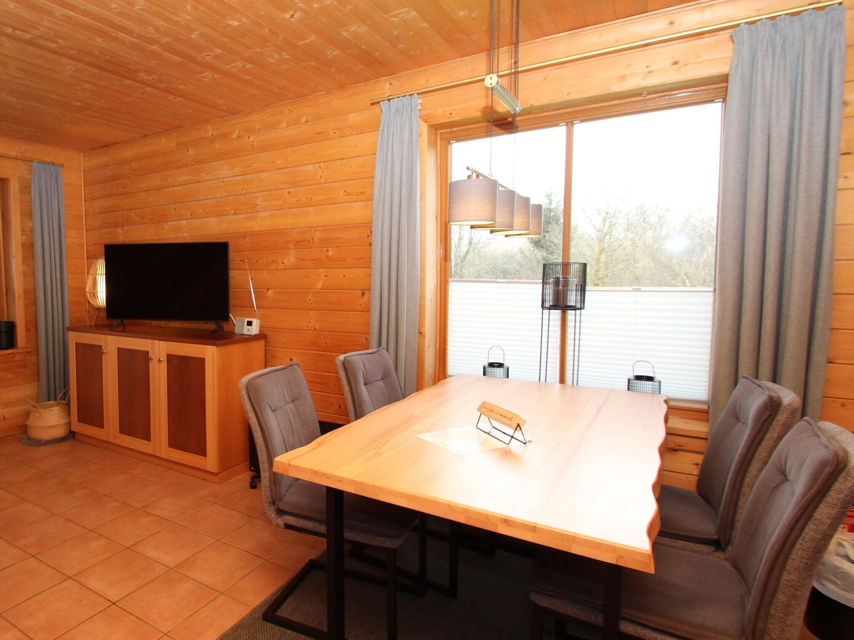 A wooden cabin interior with a dining table, chairs, a television, and a window with curtains.