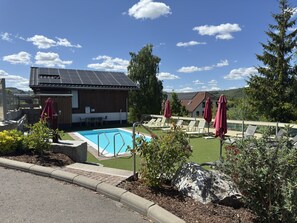 Ett poolområde med solstolar och parasoller, en byggnad med ett sluttande tak och utsikt över det omgivande landskapet.
