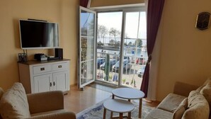 A living room with two sofas, a small table, a TV, and a sliding glass door leading to a balcony with a view of parked cars and trees. - Ferienanlage Orther Reede, Fehmarn-orth (Fehmarn)