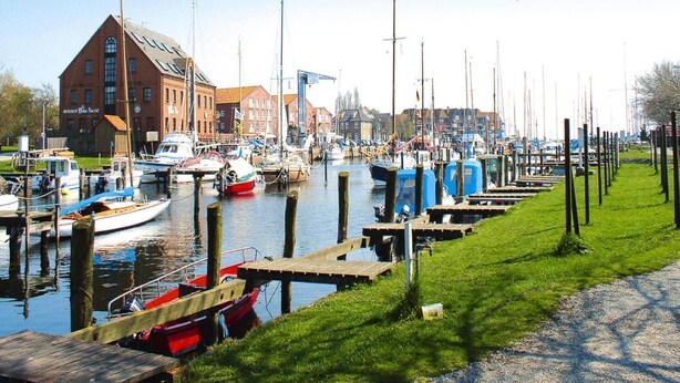 A marina with boats docked, a large brick building, and a grassy area with wooden docks. - Ferienanlage Orther Reede, Fehmarn-orth (Fehmarn)