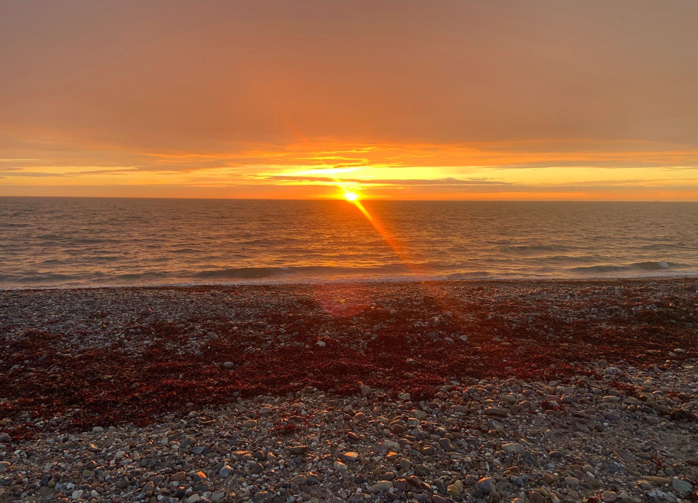 A sunset over a rocky beach with the sun visible near the horizon.