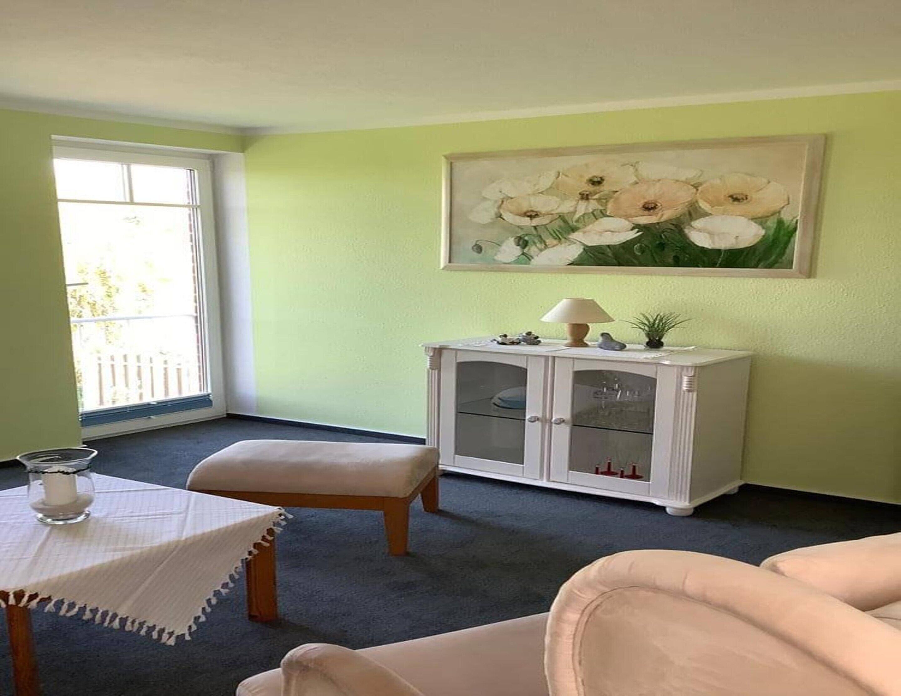 Una habitación con una pared verde, un armario blanco con puertas de vidrio, una mesa con mantel blanco y un cuadro de flores.
