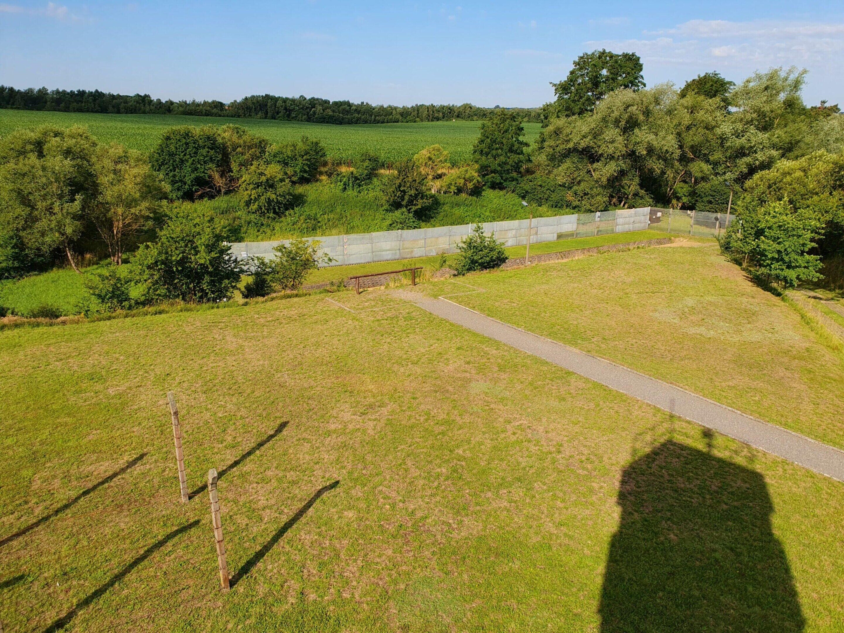 A grassy field with a paved path, trees, and a stone wall.