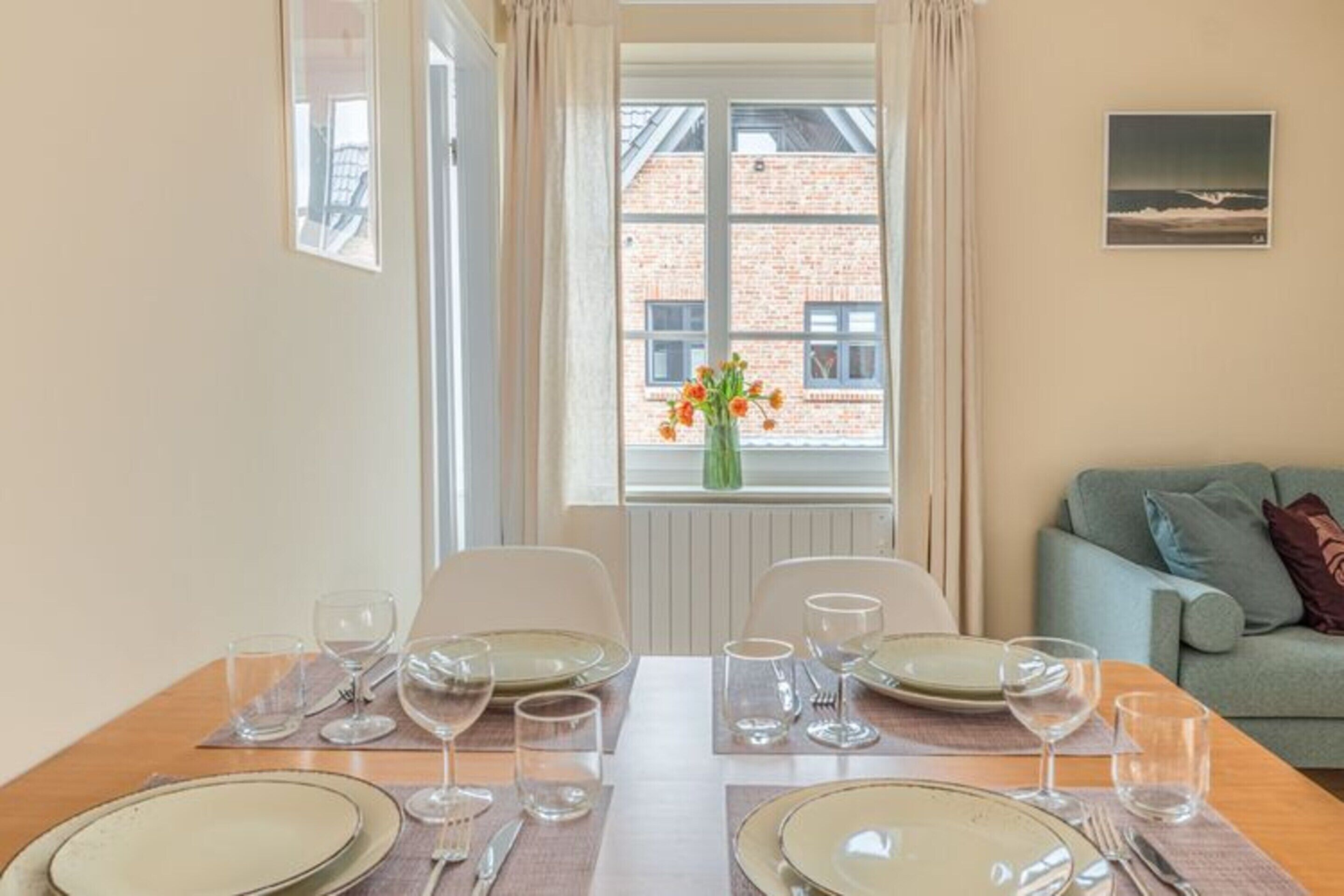 A dining area with a table set for four, chairs, a window with curtains, a vase of flowers, and a view of a brick building.
