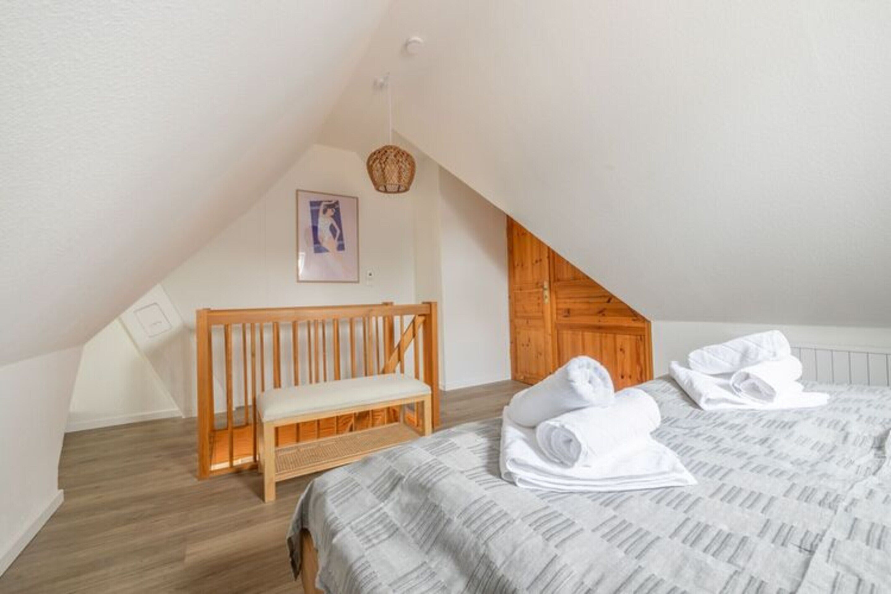 Une chambre avec un plafond en pente, un banc en bois, un lit avec des serviettes roulées et un tableau encadré au mur.
