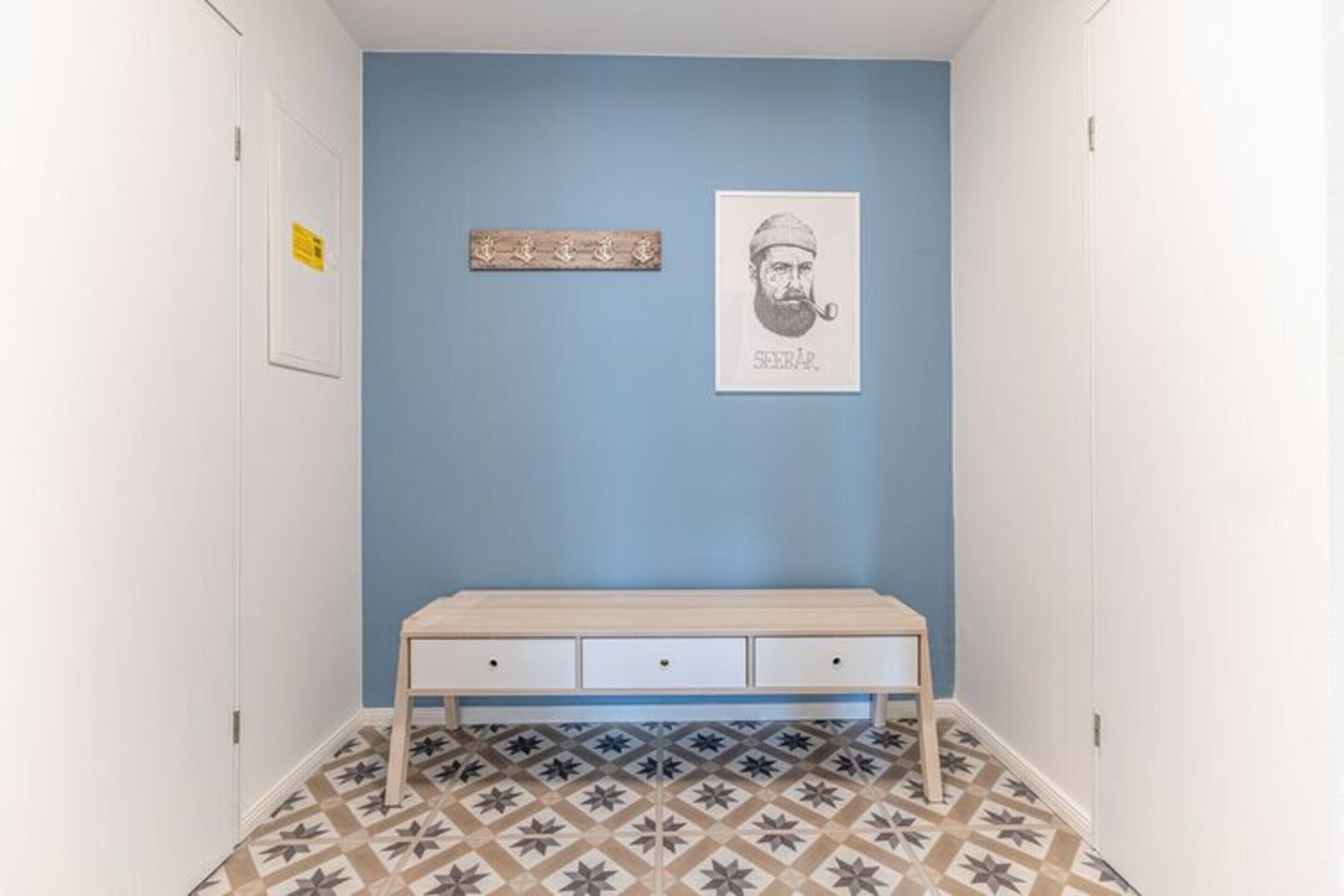 A hallway with a blue wall, a wooden bench with drawers, patterned floor tiles, and a framed picture on the wall.
