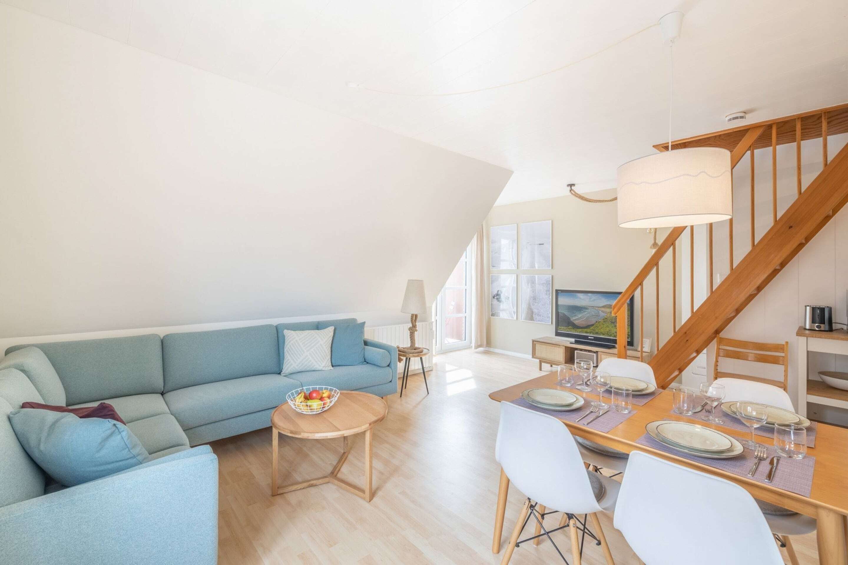 A spacious living area with a blue sofa, a dining table set for four, and a wooden staircase.