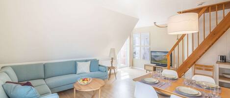 A spacious living area with a blue sofa, a dining table set for four, and a wooden staircase.