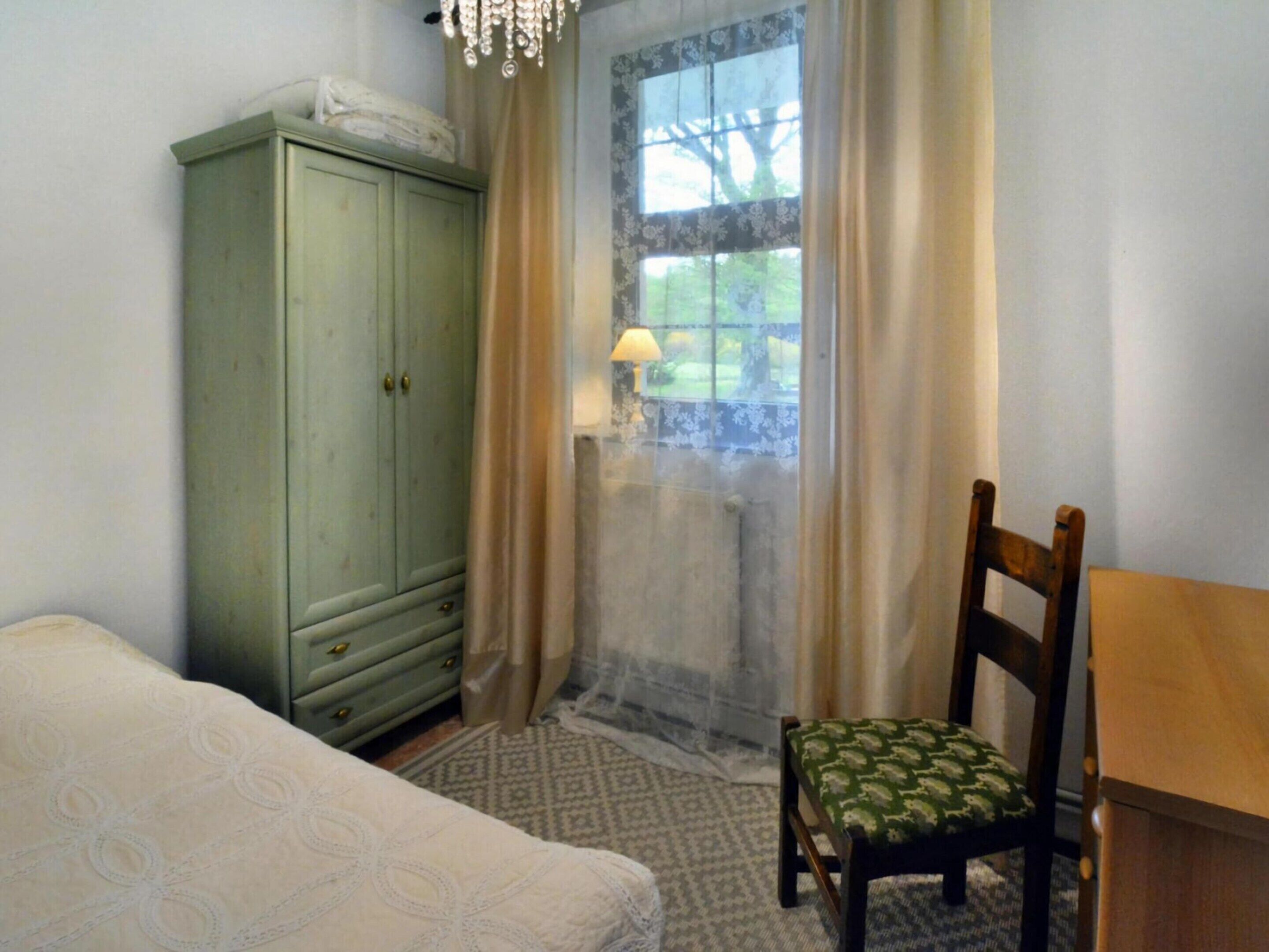Un dormitorio con cama, una silla de madera, un escritorio de madera, un armario verde y una ventana con cortinas.