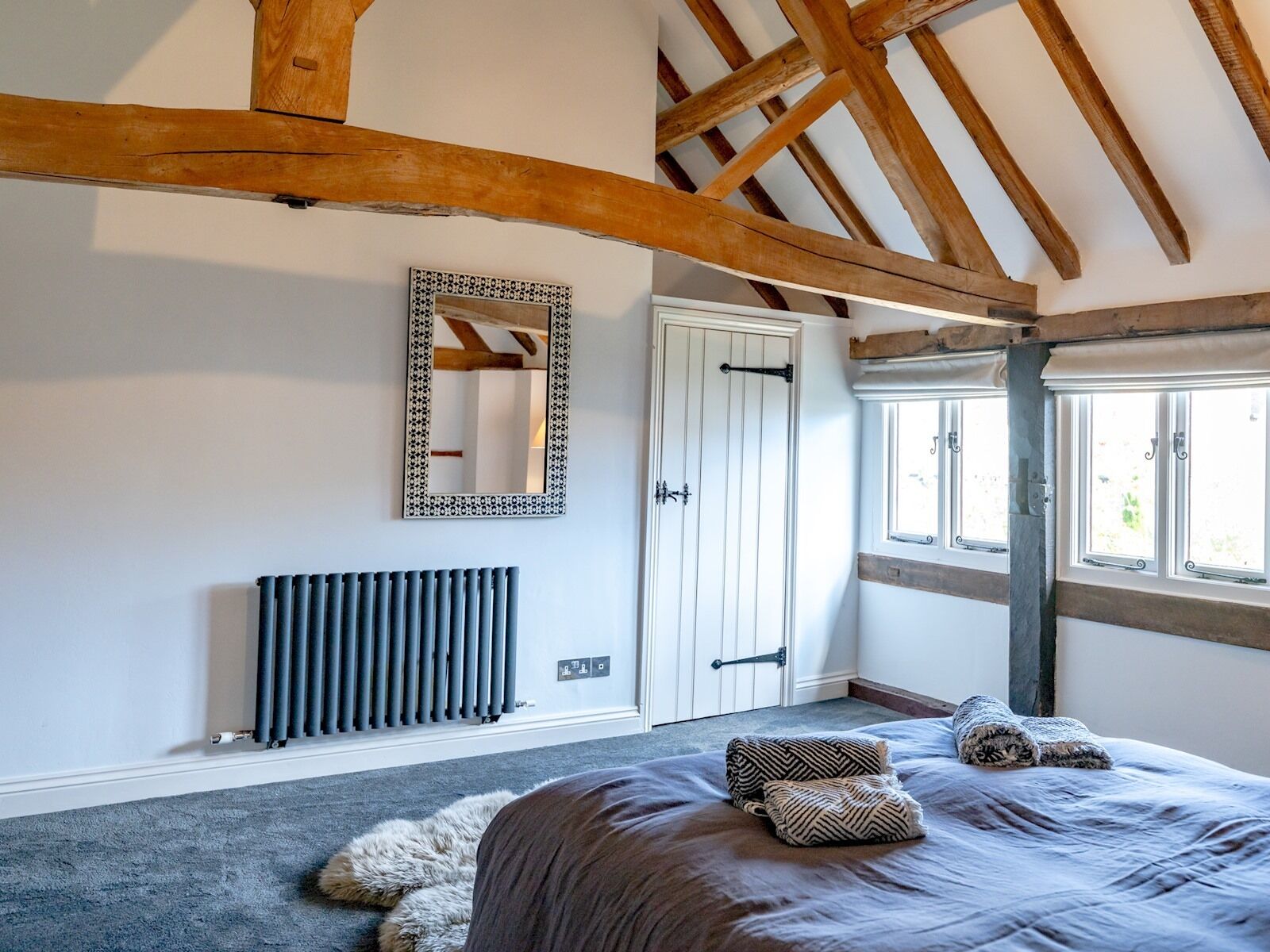 A bedroom with a large bed, a radiator, a wooden ceiling, a door, and windows.