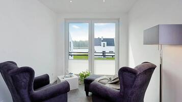 A modern living room with two dark armchairs, a small white side table, and a floor lamp. A large window offers a view of a building and a body of water.