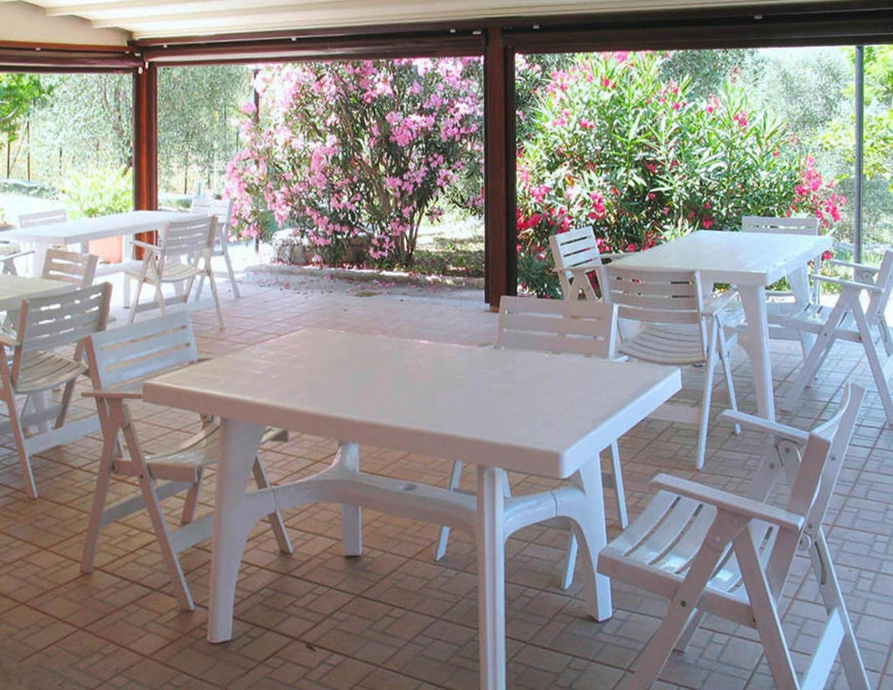 An outdoor patio area with white tables and chairs, surrounded by lush greenery and flowering plants.