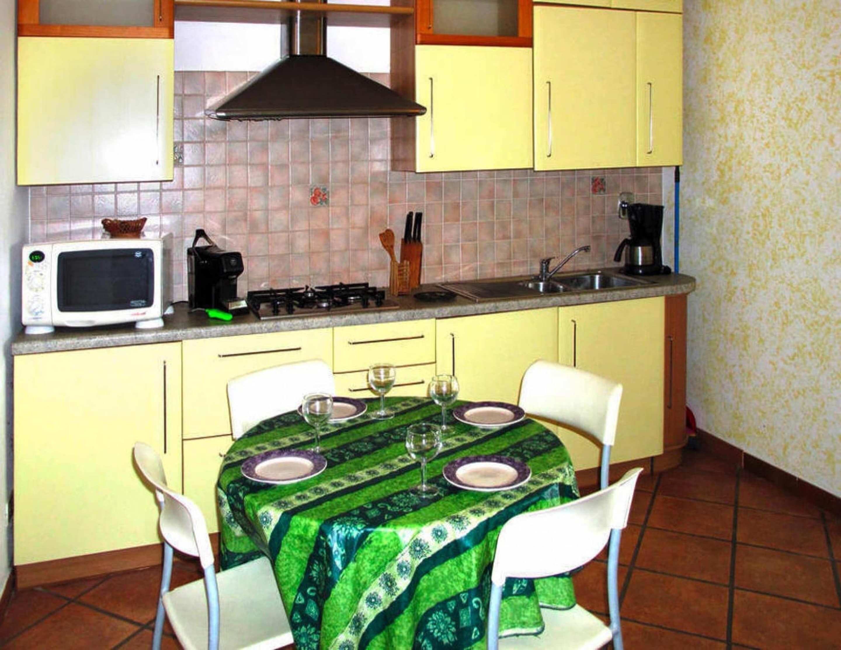 Una pequeña cocina con armarios amarillos, un microondas, una estufa y un lavabo. Hay una mesa de comedor con mantel verde y sillas blancas.