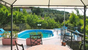 A covered outdoor area with a swimming pool, seating, and a view of the surrounding greenery and buildings. - Apartment Agriturismo Le Mimose in Imperia (Imperia)