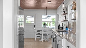 Una cocina moderna con armarios blancos, suelo de madera, un comedor con mesa y sillas blancas, y una ventana que proporciona luz natural.