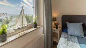 Et soveværelse med en seng, et natbord, en lampe, et vindue med udsigt til bygninger og træer, og en model af en sejlbåd på vindueskarmen.