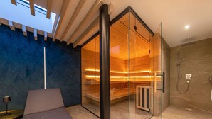 Moderni kylpyhuone, jossa on lasiseinällä erotettu sauna, suihkutila ja oleskelualue penkkeineen.