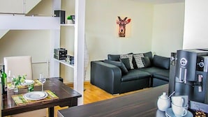 Una sala de estar moderna con una mesa de comedor marrón oscuro preparada para dos personas, un sofá negro con cojines estampados y una máquina de café a la derecha.