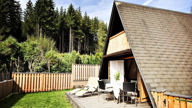 Una terraza de madera con mobiliario exterior y una pequeña mesa, rodeada por un cerco de madera y un bosque denso.