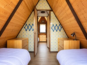 Un dormitorio abuhardillado triangular con dos camas de madera, un ventanal pequeño y un aparador de madera.