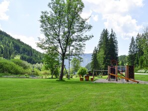 Un terrain de jeux avec des balançoires et des structures d’escalade, situé dans un parc verdoyant, avec des arbres et une montagne en arrière-plan.