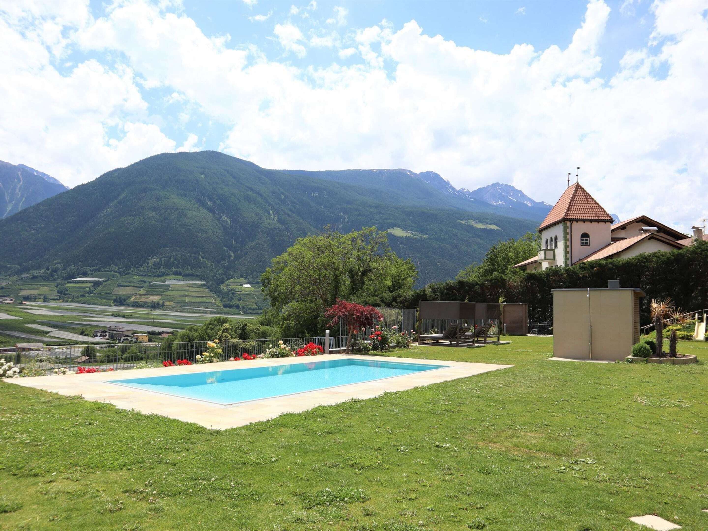 A swimming pool surrounded by a well-maintained lawn, a small building, and a church with a red-tiled roof, set against a backdrop of mountains and a clear sky.