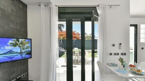 A modern living room with a flat-screen TV mounted on a dark wall, a white dining area with a table and chairs, and a sliding glass door leading to an outdoor patio with a hanging chair and a table. - Central Bungalow With Communal Pool in Maspalomas (San Bartolomé de Tirajana)