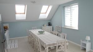 Ein Essbereich mit einem langen Tisch, weißen Stühlen und einer Schüssel auf dem Tisch. Zwei Oberlichter und ein Fenster mit Jalousien sind vorhanden.
