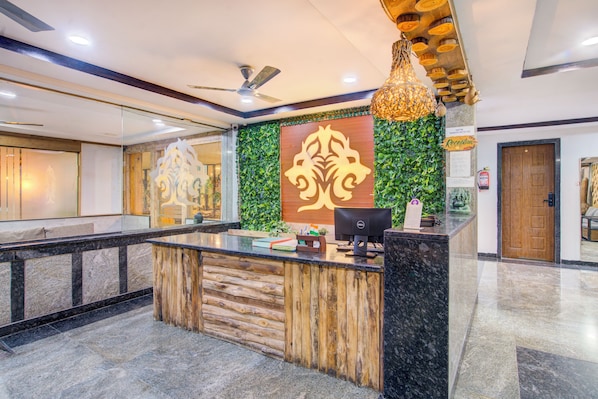 A reception area with a wooden reception desk, a green wall with a decorative emblem, a ceiling fan, and a hanging light fixture. - Hotel Loki's Forest Inn (Hyderabad)