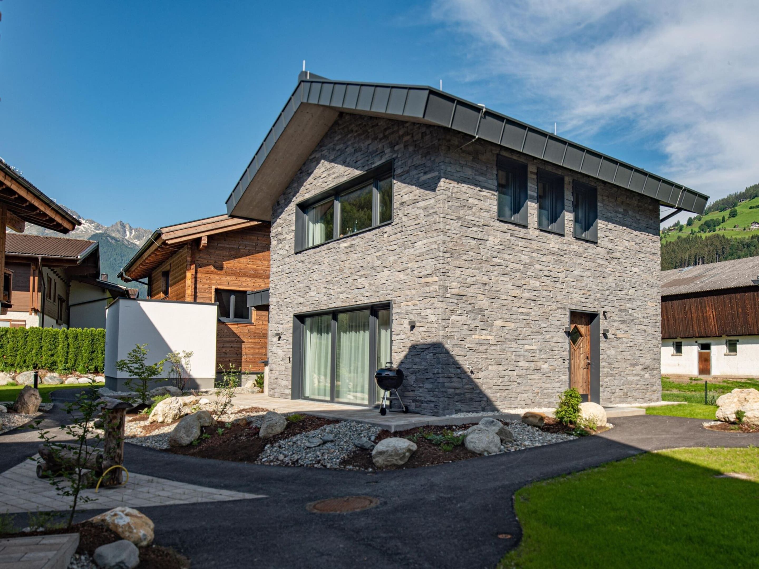 A modern two-story house with a stone facade, large windows, and a well-maintained garden with rocks and shrubs.