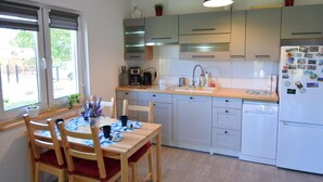 A modern kitchen with white cabinets, a wooden dining table with chairs, and a window with blinds. - Cottage in a Quiet Location, Slajszewo (Choczewo)