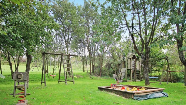 Um parquinho com balanços, um escorregador e uma casinha de madeira em uma área gramada cercada por árvores.