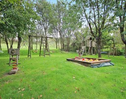 A playground with swings, a slide, and a wooden playhouse in a grassy area surrounded by trees.