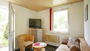 Ein gemütlicher Wohnbereich mit Sofa, Sessel, kleinem Tisch, Fernseher und einem Fenster mit Blick auf Grünflächen.