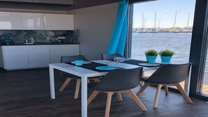 Un espace repas moderne avec une table blanche et des chaises noires, une cuisine avec des meubles blancs et une vue sur l’eau où l’on peut apercevoir des voiliers.