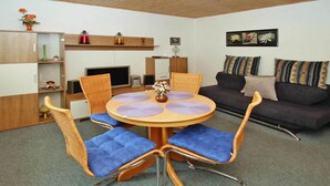 A compact living space with a round wooden dining table, blue cushioned chairs, a dark sofa with patterned cushions, and a small kitchenette area. - Ferienhaus, Plau am See (Plau am See)