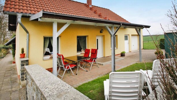 A yellow building with a red-tiled roof, a covered patio with red and white chairs, and a stone wall in the foreground. - Ferienhaus, Plau am See (Plau am See)