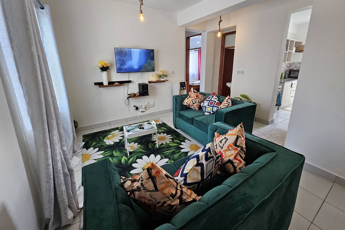 A modern living room with a flat-screen TV, green sofas, a coffee table, and a decorative rug.