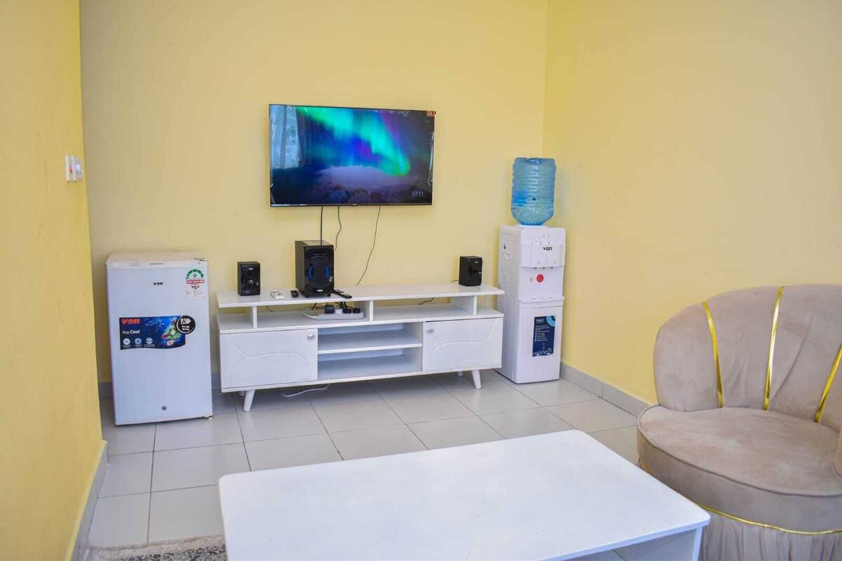 A living room with a flat-screen TV mounted on the wall, a white entertainment unit with a water dispenser, a small refrigerator, and a beige armchair.