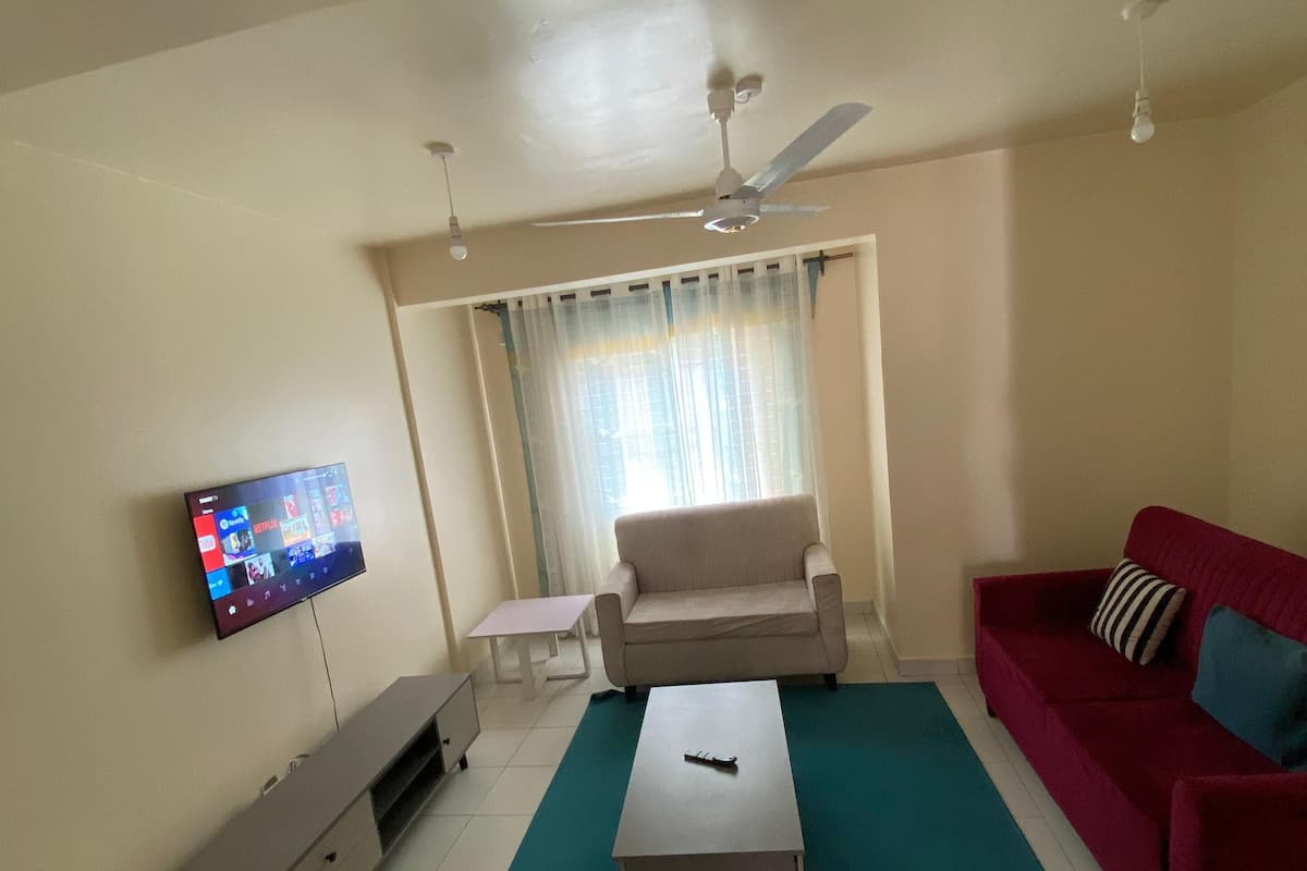 A living room with a TV, a red sofa, a beige sofa, a coffee table, a ceiling fan, and a window with curtains.