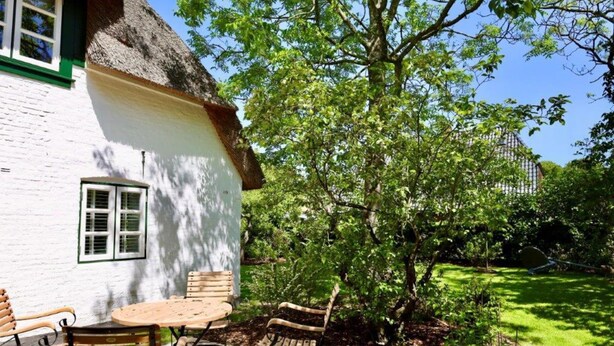 Un bâtiment blanc avec un toit de chaume, une table en bois et deux chaises en bois, le tout situé dans un jardin.