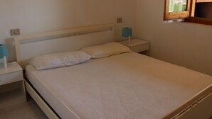 Ein Zimmer mit einem Einzelbett, einem weißen Kopfteil, einem Nachttisch mit Lampe und einem Fenster mit Blick ins Freie.