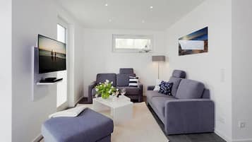A modern living room with a flat-screen TV, two grey sofas, a white coffee table, and a large window.