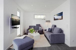 A modern living room with a flat-screen TV, two grey sofas, a white coffee table, and a large window.