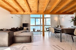 Ein modernes Wohnzimmer mit einer großen Eckcouch, einem Essbereich mit Tisch und Stühlen und einem großen Fenster mit Blick auf die Berge.