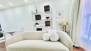 A living room with a beige sofa, a glass coffee table, and a shelving unit with books and decorative items.