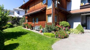 Une maison en bois à deux étages, dotée d’un balcon, d’un jardin avec différentes plantes et fleurs, et d’un banc.