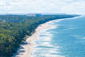 A coastal view with a beach, forested area, and a large cruise ship docked at a pier. - Cottage in Pustkowo Near Sandy Beach (Rewal)