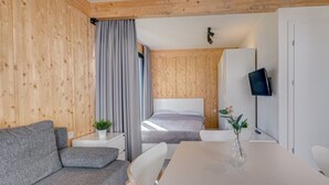 Une chambre moderne avec un canapé gris, une table blanche ornée d’un vase, un lit avec une tête de lit grise et un téléviseur fixé au mur.