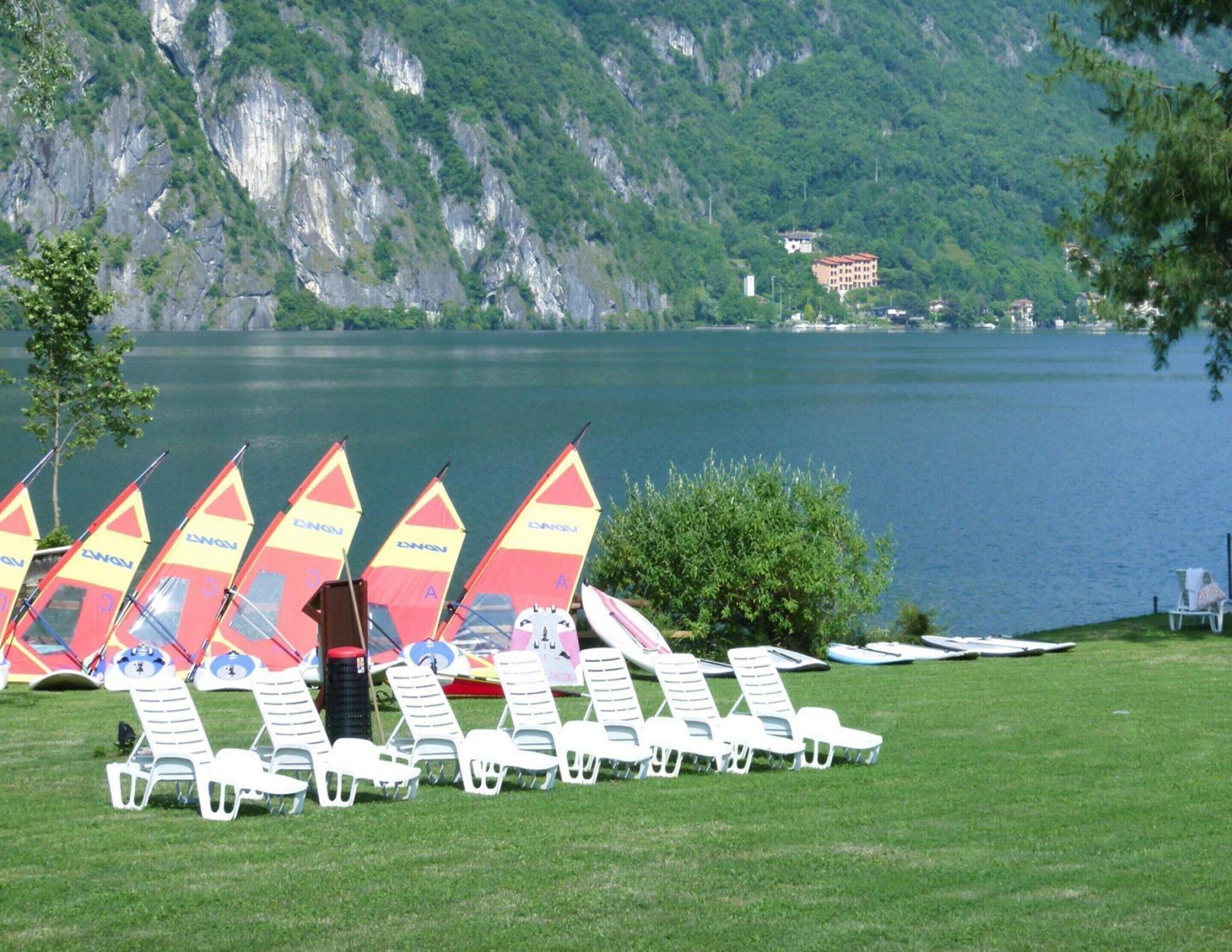 A lakeside area with white lounge chairs and several sailboats with red and yellow sails.